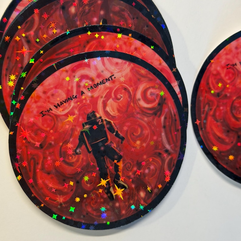 May include: A stack of round, red holographic stickers with a black border. Each sticker features an astronaut floating in space with the text "I'M HAVING A MOMENT." The stickers have a swirling red background and colorful star accents.