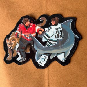 Könnte beinhalten: Ein Aufkleber mit zwei Eishockeyspielern in Trikots, eines rot und eines weiß, in einer humorvollen Szene mit einem Puma und einem Hai. Der Aufkleber hat einen schwarzen Rand und ist vor einem braunen Hintergrund platziert.