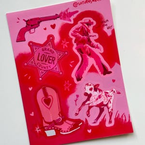 May include: A set of pink and red stickers featuring a cowboy, a boot, a calf, a gun, and a sheriff's star with the text "Grand Lover County". The stickers are on a pink background with the text "@saturnswar.jpg" in the top right corner.