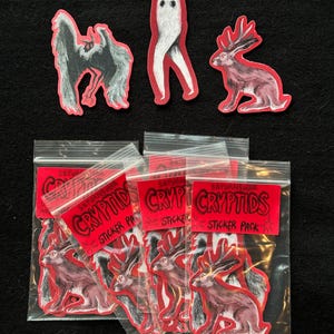 Puede incluir: Una colección de pegatinas con ilustraciones de críptidos. Las pegatinas incluyen una criatura parecida a un murciélago, un fantasma y un jackalope. Se ven varios paquetes de pegatinas, cada uno con una etiqueta roja que dice "CRYPTIDS STICKER PACK".