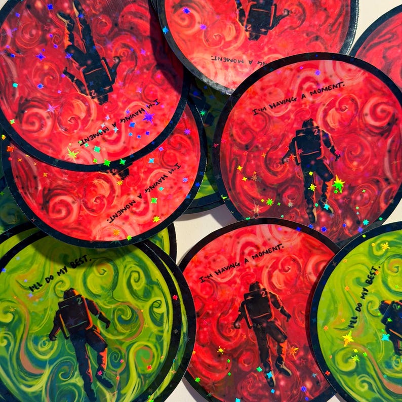 May include: A collection of round stickers with a space theme. The stickers feature an astronaut silhouette against a swirling red or green background. Text on the stickers reads "I'M HAVING A MOMENT" and "I'LL DO MY BEST."