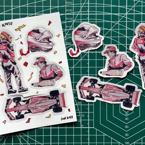 May include: A sticker sheet with illustrations of Fernando Alonso, depicting him in his racing gear, including a helmet, racing suit, and Formula 1 car. The stickers are in red, white, and black, with the name "Fernando Alonso" at the top.