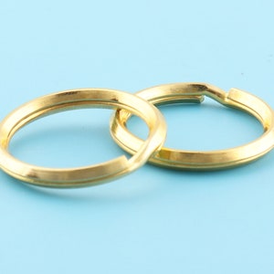 May include: Two gold-colored split key rings, interlocked, against a light blue background. The rings are circular with a flat, slightly beveled edge. The metal has a matte finish, and the rings are open for attaching keys or other items.
