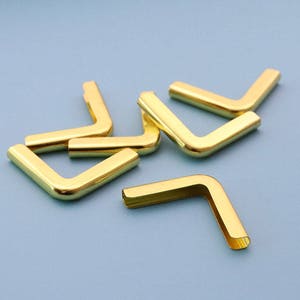 May include: A group of eight gold metal corner protectors. The corner protectors are L-shaped and have a smooth, polished finish.