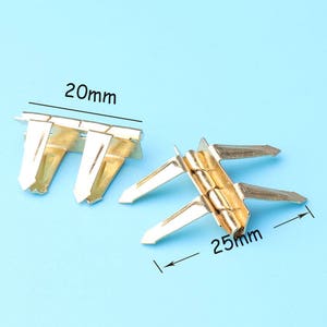 Puede incluir: Dos bisagras de metal de color dorado, una abierta y otra cerrada. La bisagra abierta mide 25 mm de largo y la bisagra cerrada mide 20 mm de largo.