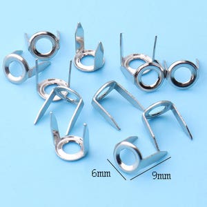 May include: A collection of silver-colored metal rivets with a circular head and three prongs. The rivets are 9mm wide and 6mm high, displayed on a light blue background. These fasteners are suitable for various craft and design projects.