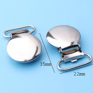 Pode incluir: Dois clipes de metal prateado para suspensórios. Cada clipe tem formato de disco plano e arredondado, fecho articulado e laço retangular. Um tem 3,5 cm de comprimento e o outro 2,2 cm de largura.