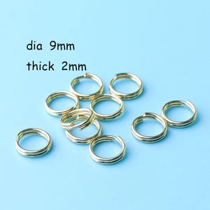 Puede incluir: Primer plano de nueve anillos de salto dorados, cada uno con un diámetro de 9 mm y un grosor de 2 mm. Los anillos están dispuestos en una pila suelta sobre un fondo azul claro.