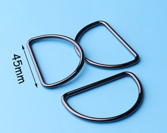 Puede incluir: Tres anillos en D de metal negro, cada uno con un diámetro de 45 mm. Los anillos están dispuestos en una pila suelta sobre un fondo azul claro.