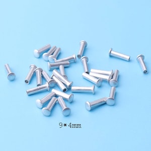 Puede incluir: Primer plano de un montón de remaches de metal plateado, cada uno mide 9 mm por 4 mm. Los remaches están esparcidos sobre un fondo azul claro.