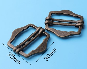 May include: Two antique gold-toned metal slider buckles, one measuring 35mm by 30mm. The buckles have a rectangular shape with rounded corners and two parallel slots for a strap.