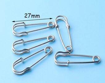 May include: Six silver safety pins, each measuring 27 mm in length, are arranged on a light blue background.