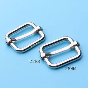 Pode incluir: Duas fivelas deslizantes de metal prateado, uma medindo 22 mm e a outra 25 mm. As fivelas são projetadas para uso com correias ou tiras.