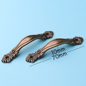 Puede incluir: Dos tiradores de cajón de bronce de estilo antiguo con detalles ornamentados. Los tiradores miden 70 mm de largo y 10 mm de ancho.