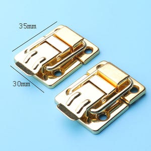 Puede incluir: Dos cierres metálicos dorados, cada uno con una base rectangular y un cierre con bisagras. Los cierres miden 35 mm x 30 mm. El fondo es de color azul claro liso.