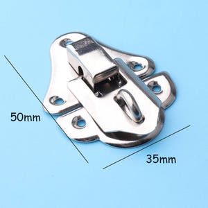 Puede incluir: Pestillo de metal plateado de forma rectangular de 50 mm x 35 mm. El pestillo tiene un mecanismo de resorte y un pequeño bucle para un candado.