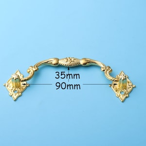 Puede incluir: Tirador de cajón de metal dorado con un diseño decorativo. El tirador mide 35 mm por 90 mm.