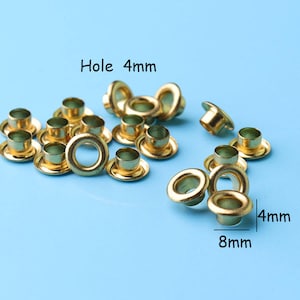 Puede incluir: Primer plano de 17 ojales metálicos de color dorado con un agujero de 4 mm y un diámetro exterior de 8 mm. Agujero 4 mm.