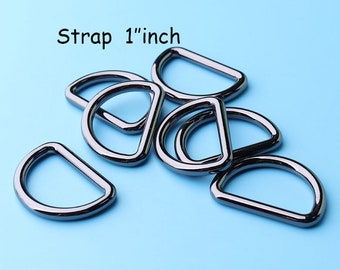 Puede incluir: Primer plano de un montón de anillas en D de metal negro, cada una con un diámetro de 2,5 cm. Las anillas están dispuestas de forma aleatoria sobre un fondo azul claro. El texto "Strap 1" inch" es visible en la parte superior de la imagen.