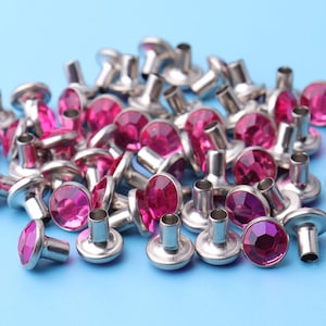 May include: A pile of silver metal rivets with pink rhinestones. The rivets have a flat head and a pointed end.