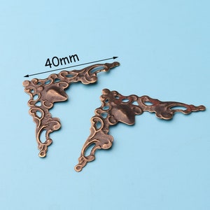 May include: Two antique gold-toned metal corner brackets with a decorative floral design. The brackets measure 40mm in length.