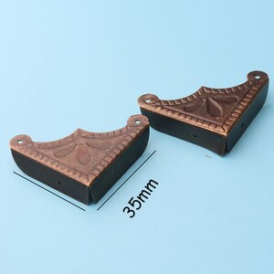 Może przedstawiać: Dwa antyczne mosiężne narożniki z ozdobnym wzorem. Każdy narożnik ma 8,9 cm długości.