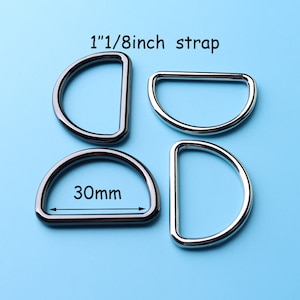 Op de afbeelding: Vier zilverkleurige D-ringen, twee met een maat van 1 1/8 inch en twee met een maat van 30 mm, op een lichtblauwe achtergrond.