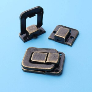 May include: Three antique bronze-colored metal latches on a light blue background. The latches are rectangular with a hinged design, featuring a clasp and a catch. The metal has a distressed finish, suggesting an aged appearance, and is suitable for boxes or chests.