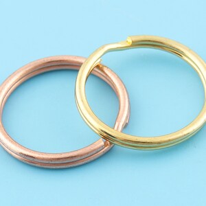 May include: Two metal key rings, one in a rose gold color and the other in gold, are displayed against a light blue background. The rings are circular and have a split design for easy attachment of keys or other items.