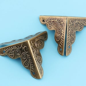 Könnte beinhalten: Zwei verzierte Eckenschutz aus Metall mit einem dekorativen Blumenmuster. Die Schutzecken sind aus goldfarbenem Metall und haben einen Vintage-Look.