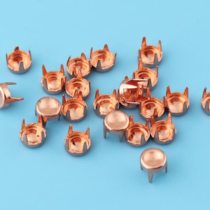May include: A group of 17 copper-colored metal rivets with four prongs on the bottom. The rivets are scattered on a light blue surface.