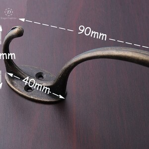 May include: Antique bronze coat hook with a curved design. The hook measures 90mm long, 40mm wide, and 35mm high.