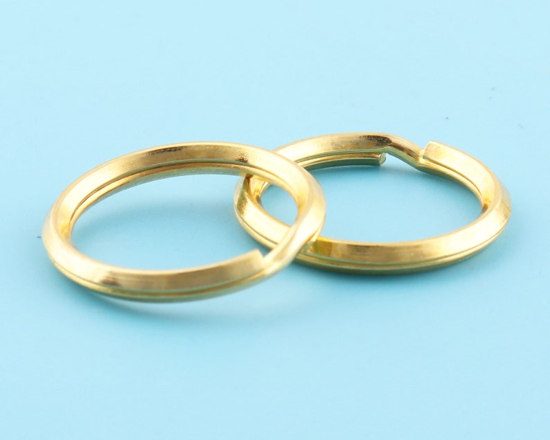 May include: Two gold-colored split key rings, interlocked, against a light blue background. The rings are circular with a flat, slightly beveled edge. The metal has a matte finish, and the rings are open for attaching keys or other items.