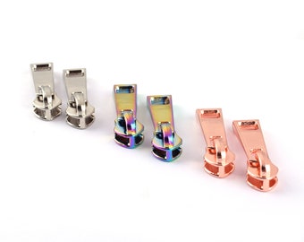 May include: A collection of zipper pulls in three different metallic finishes: silver, iridescent rainbow, and rose gold. The pulls are designed with a rectangular top and a rounded, double-layered base.