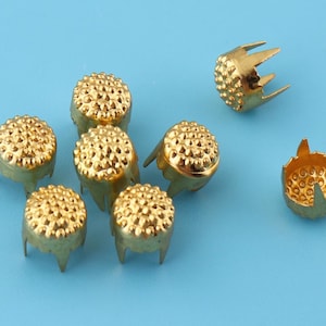 May include: Eight gold-colored metal studs with a textured surface and three prongs on the bottom. The studs are arranged in a cluster on a light blue background.