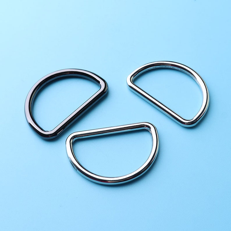 May include: Three metal D-rings, two silver and one black, on a light blue background.