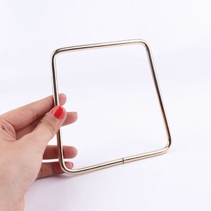 May include: Gold-toned metal handle for a bag or purse. The handle is square shaped and has a smooth finish.