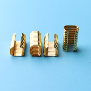 May include: Four gold-colored metal clips, each with a rectangular shape and a ribbed texture. The clips are arranged in a row on a light blue background.