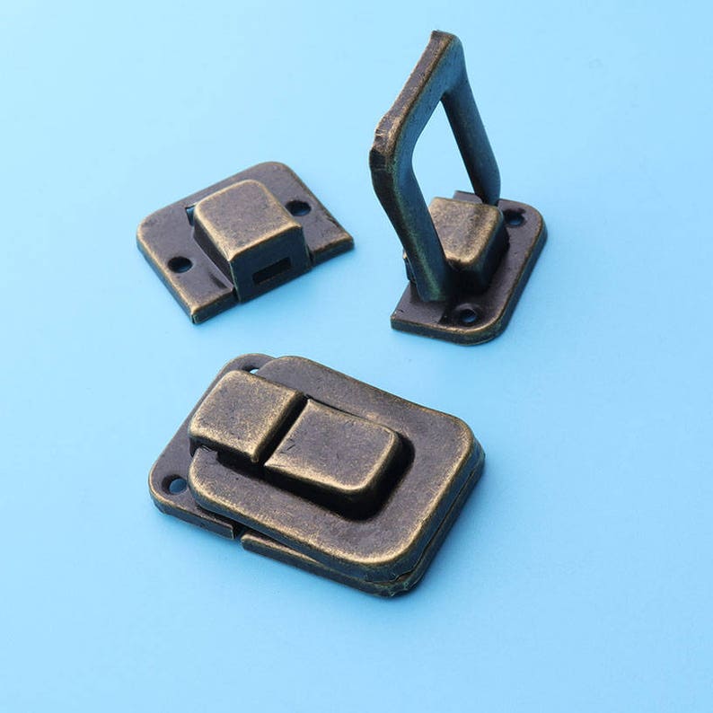 May include: Three antique bronze-colored latches on a light blue background. One latch is open, showing a handle, while the other two are closed. Each latch has a rectangular base with screw holes.