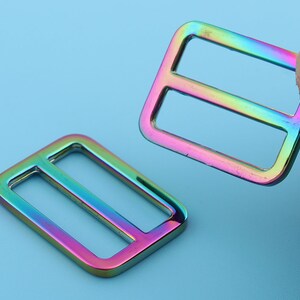 May include: Two iridescent rainbow-colored metal slider buckles. The buckles are rectangular with a single horizontal slot in the center.