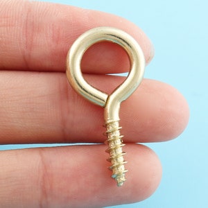 Puede incluir: Un gancho de ojo de tornillo de color dorado con un bucle redondeado. El gancho se sostiene en la mano de una persona.