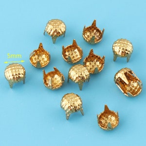 May include: A close-up of 10 gold-colored metal studs with a domed top and three prongs on the bottom. The studs are 5mm in diameter.