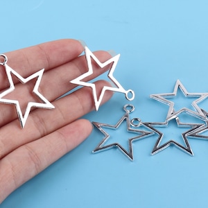 May include: Six silver metal star-shaped charms with open centers and jump rings. The charms are arranged on a blue surface.