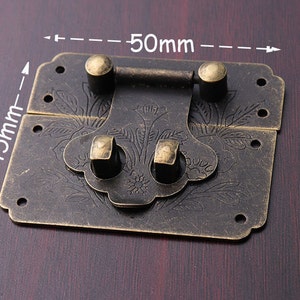 Może przedstawiać: Antyczne mosiężne metalowe zamknięcie, 45 mm x 50 mm, z kwiatowym grawerunkiem. Zamknięcie posiada centralny obrotowy drążek i dwa zakrzywione zatrzaski. Metal ma postarzaną powierzchnię. Idealne do pudełek w stylu vintage.