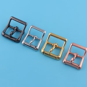 Puede incluir: Cuatro hebillas metálicas en diferentes acabados: negro, plateado, dorado y oro rosa. Cada hebilla tiene un marco rectangular con una barra central y un pasador para cerrar. Las hebillas están dispuestas sobre un fondo azul claro.