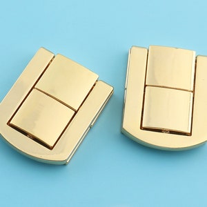 Puede incluir: Dos cierres metálicos de color dorado con forma rectangular y diseño de bisagra. Los cierres tienen un acabado liso y pulido.