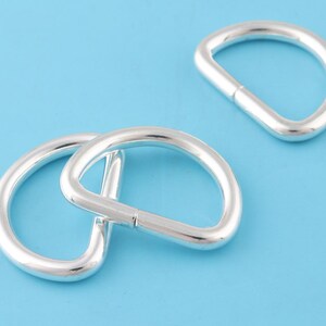 Puede incluir: Tres anillos en D de metal plateado, también conocidos como llaveros, se muestran sobre un fondo azul claro. Los anillos tienen un diámetro de aproximadamente 2,5 cm.