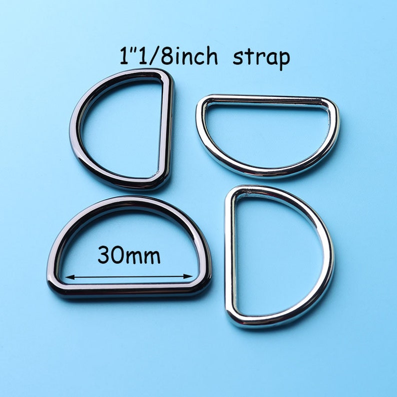 May include: Four silver-toned D-rings, two measuring 1 1/8 inch and two measuring 30mm, on a light blue background.