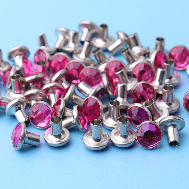 May include: A pile of silver metal rivets with pink rhinestones. The rivets have a flat head and a pointed end.