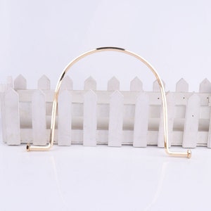 May include: Gold metal handbag handle with a rounded top and two straight ends. The handle is on a white picket fence background.